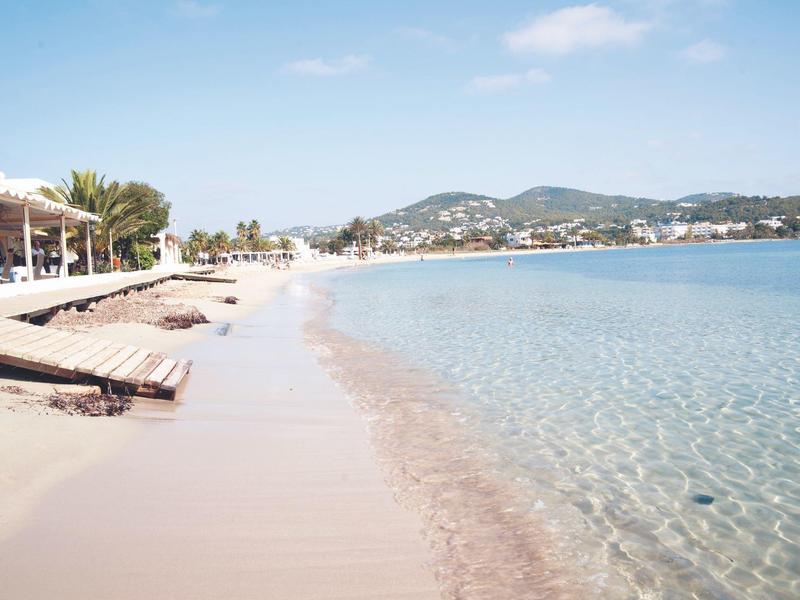 A calm sandy beach with clear water and sun loungers along the shore under a blue sky.