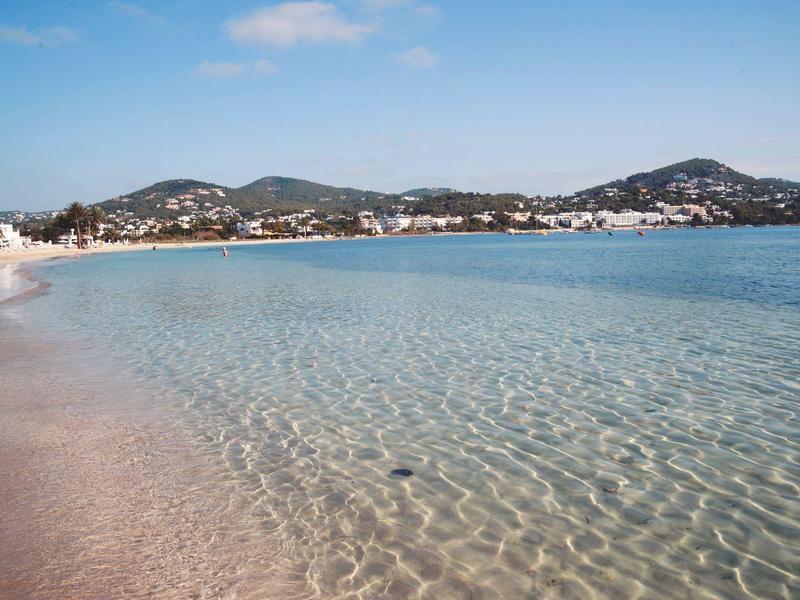 Clear, calm sea water at a beach with mountains and houses in the background.