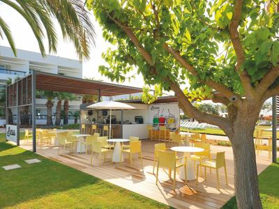 Terrazza dell'hotel con tavoli bianchi, ombrelloni, alberi e prato verde.