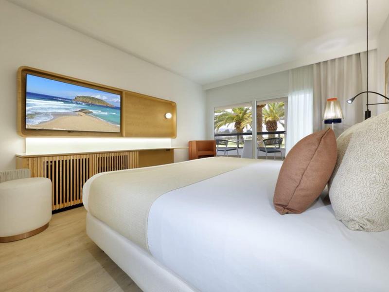 Camera d'hotel luminosa con grande letto, vista su palme, arredi moderni e pareti chiare.