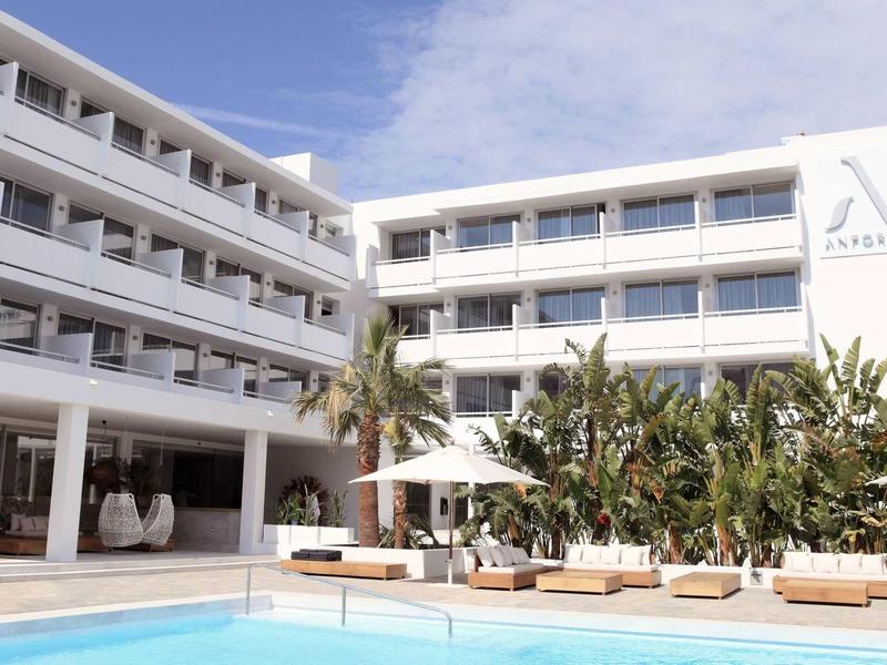 Modern hotel with white facade, pool, and tropical plants under a blue sky.