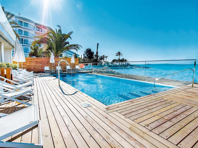Helles Schwimmbad mit Holzterrasse und Liegestühlen am Meer an sonnigem, klarem Tag.