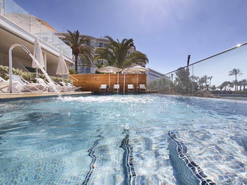 Tunnelblick auf einen klaren Pool mit Liegestühlen, Palmen und hellblauem Himmel im Hintergrund.