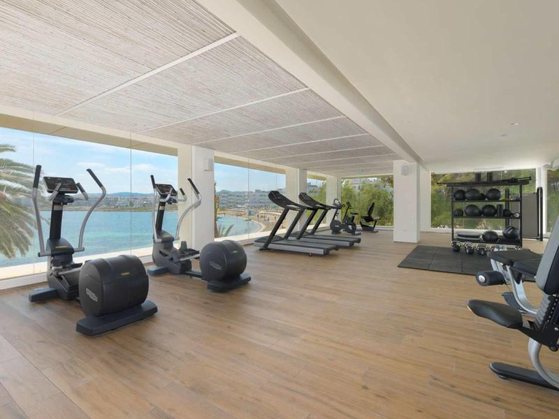 Palestra moderna con attrezzature cardio e vista panoramica sul mare e palme.