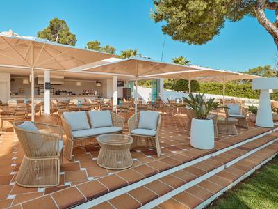 Terrasse avec mobilier en osier et parasols devant un bâtiment hôtelier sous un ciel bleu clair.