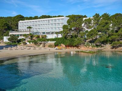 Hôtel moderne au bord d'une plage calme avec une eau claire et de nombreux arbres en arrière-plan.
