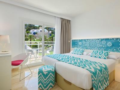 Chambre d'hôtel lumineuse avec lit double, linge de lit bleu et blanc, et balcon donnant sur des collines verdoyantes.