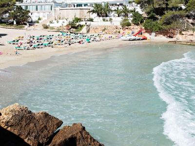 Strand mit weißen Sand und klarem Wasser vor einem mehrstöckigen Hotelgebäude und Bäumen
