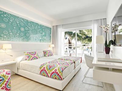 Modernes Hotelzimmer mit Doppelbett, bunten Decken und Blick auf tropische Pflanzen.