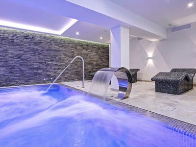 Modernes Hallenbad mit beleuchteter Wasserfallfunktion und Liegestühlen vor steinerner Wand.