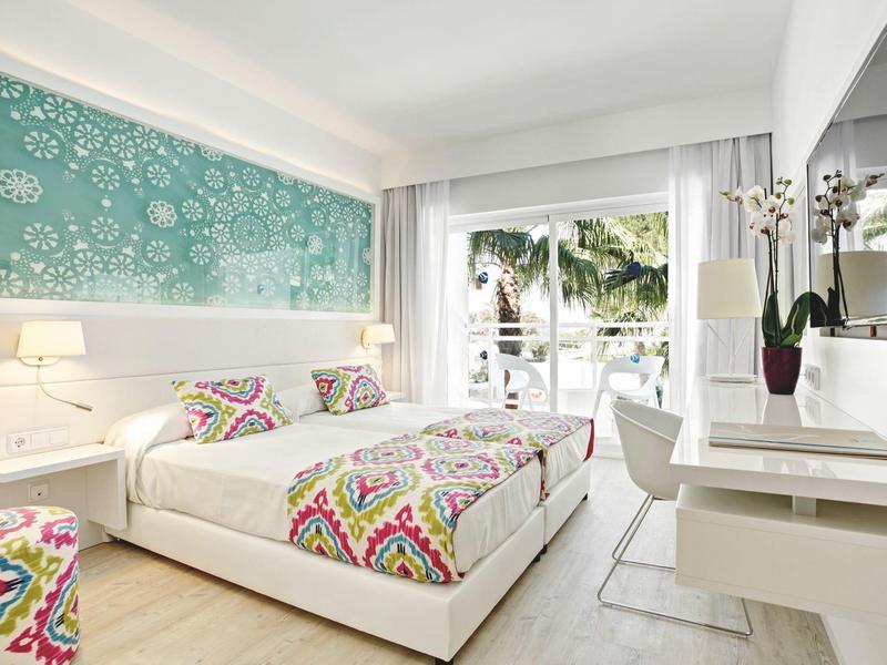 Modernes Hotelzimmer mit Doppelbett, bunten Decken und Blick auf tropische Pflanzen.