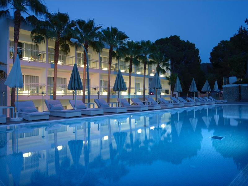 Beleuchteter Hotelpool mit Liegestühlen und Sonnenschirmen bei Nacht.