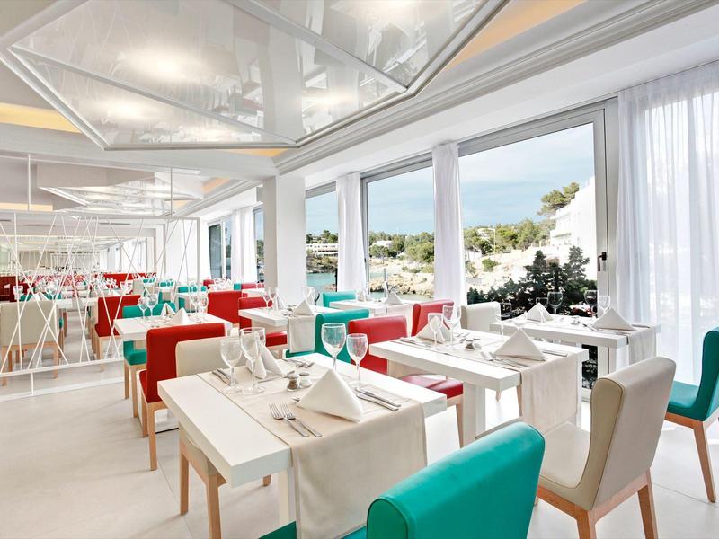 Modernes Restaurant mit weißen Tischen, bunten Stühlen und großen Fenstern mit Blick auf Landschaft.