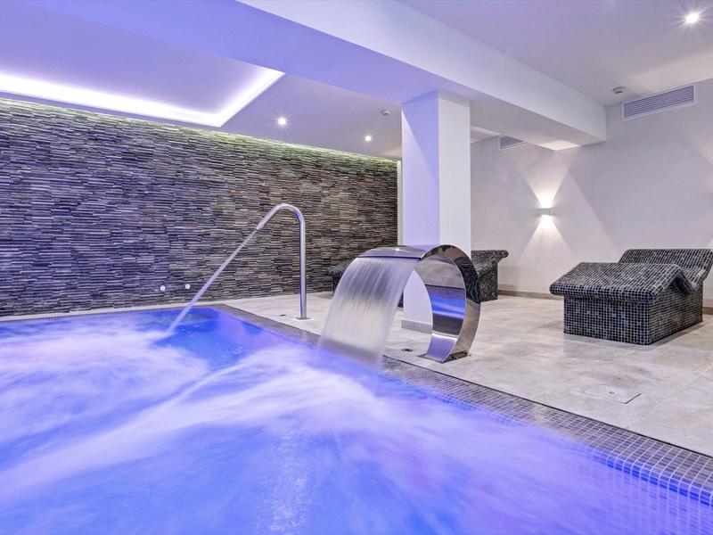 Modernes Hallenbad mit beleuchteter Wasserfallfunktion und Liegestühlen vor steinerner Wand.