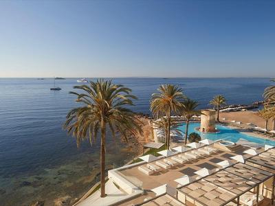Vista su una terrazza di un hotel costiero con palme e piscina sul mare sotto un cielo sereno.