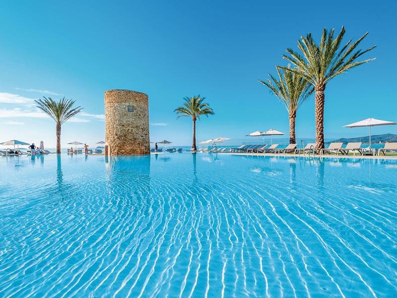 Acqua blu chiara in una piscina a sfioro con palme e ombrelloni sotto un cielo limpido.