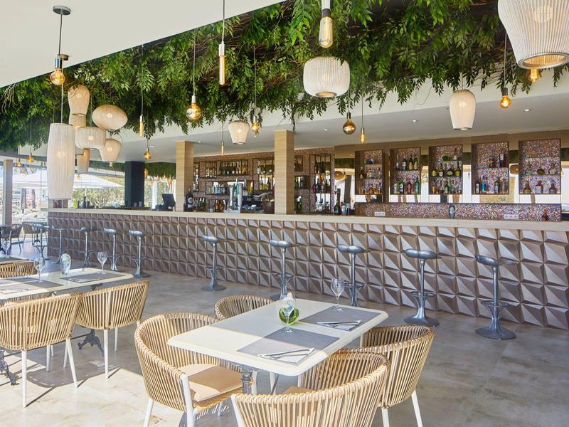 Bar d'hotel moderna con lampade, piante verdi e aree con tavoli e sedie.