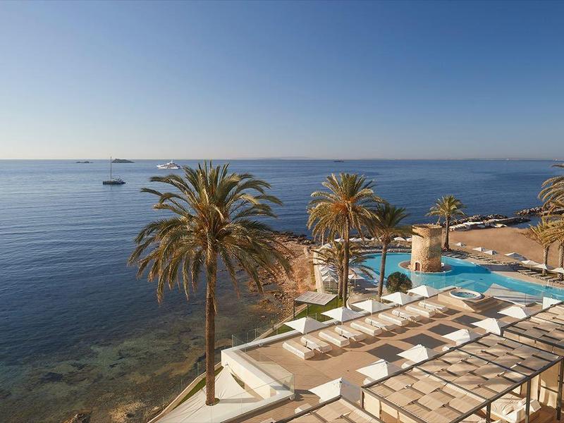 Vista su una terrazza di un hotel costiero con palme e piscina sul mare sotto un cielo sereno.