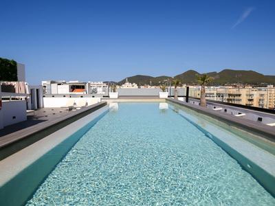 Schwimmbad auf Dach mit klarem Wasser, umgeben von Gebäuden und Bergen unter blauem Himmel.