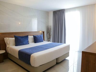 Hell eingerichtetes Schlafzimmer mit großem Bett, blauen Kissen, Vorhängen und Holzmöbeln.