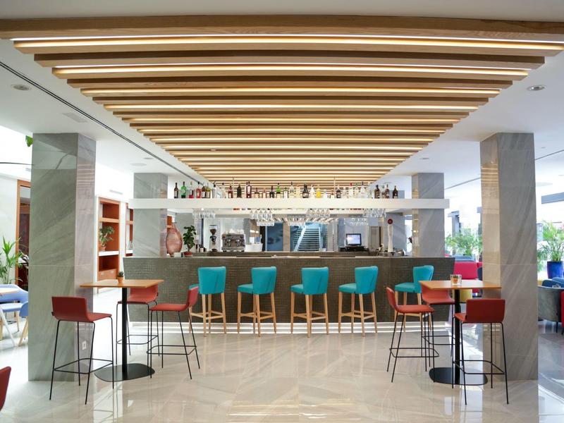 Moderner Barraum mit Holzdecke, hellen Stühlen und Theke, offene Regale und große Fenster.