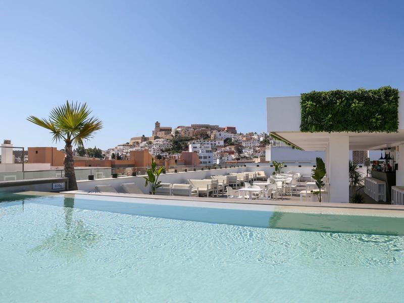 Infinity-Pool mit klarem Wasser, Palmen und weißer Stadt auf Hügel im Hintergrund unter blauem Himmel.