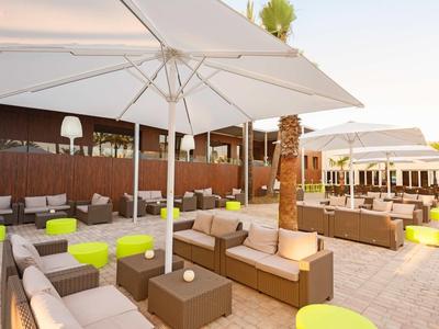 Nowoczesna przestrzeń na zewnątrz z beżowymi sofami, białymi parasolami i jasnymi zielonymi stołkami na brukowanej podłodze.