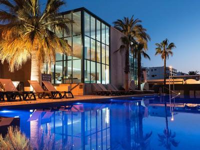 Moderner Pool am Abend mit beleuchteter Terrasse, Liegestühlen und Palmen unter blauem Himmel.