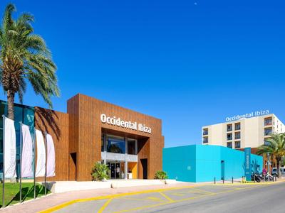 Modernes Hotelfrontgebäude mit brauner Fassade, blauem Anbau, Palmen und klarem blauem Himmel.