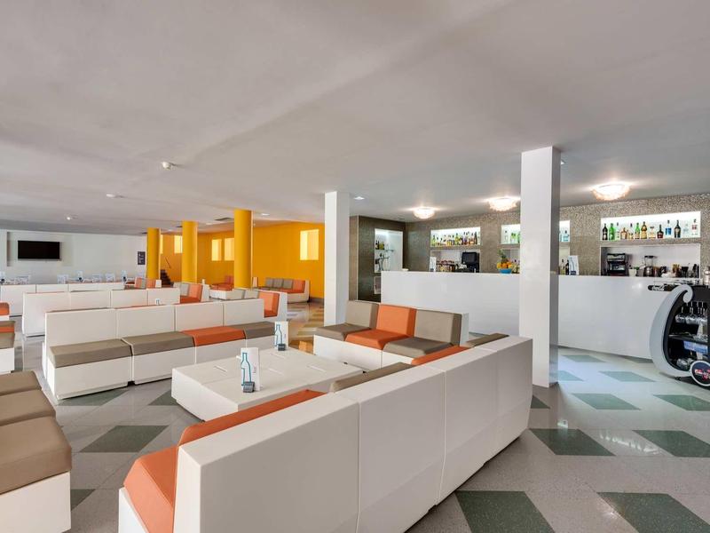 Moderner, heller Lounge-Bereich mit weißen und orangen Sesseln, Tischen und eingelassenen Deckenlichtern.