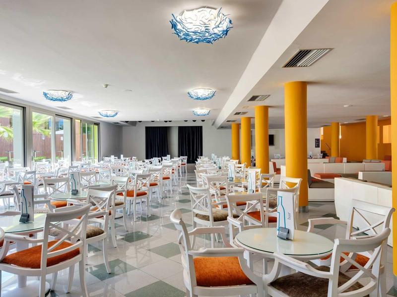 Hell gestalteter Speisesaal mit weißen Stühlen, orangen Polstern und großen Fenstern an der Seite.