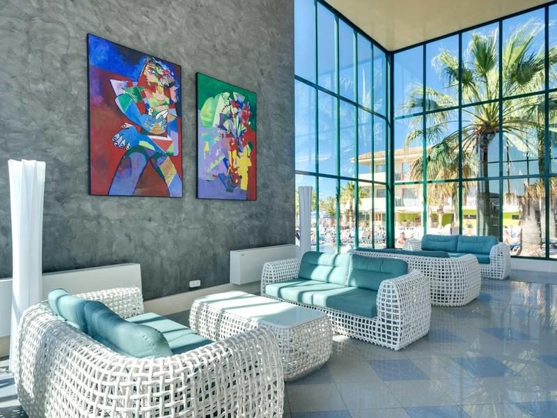 Moderner Wohnraum mit großen Fenstern, weißen Korbsesseln, blauen Kissen und bunter Wandkunst.