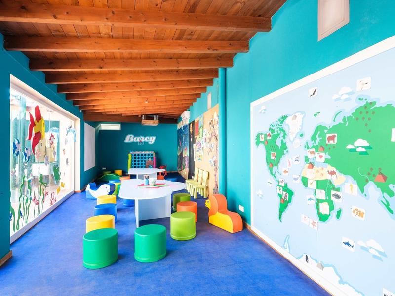 Heller Raum mit blauem Boden, bunter Sitzmöbel, Holzbalkendecke und großen Weltkarten an den Wänden.