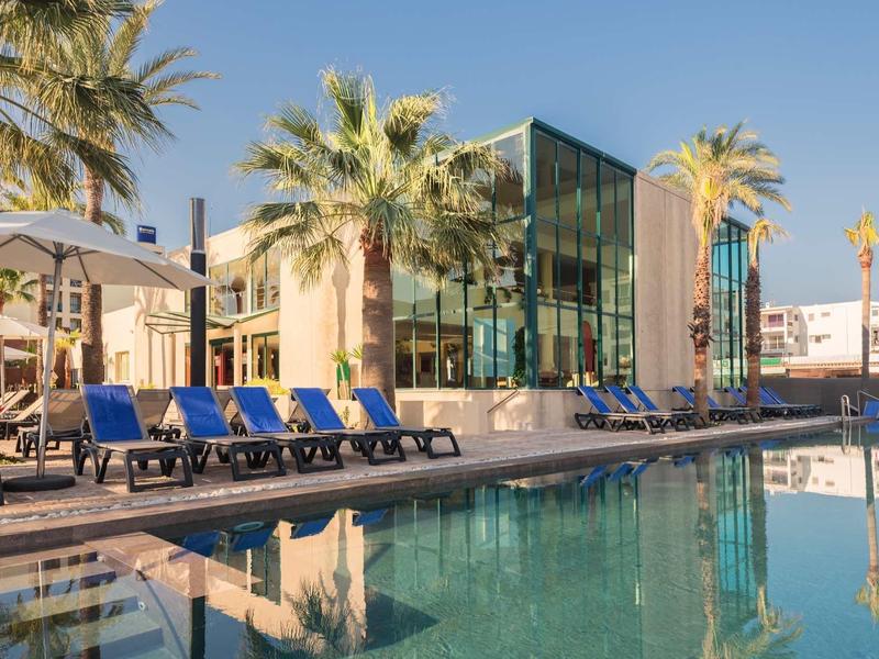 Moderner Hotelpool mit blauen Liegestühlen, Sonnenschirmen und Palmen an sonnigem Himmel.