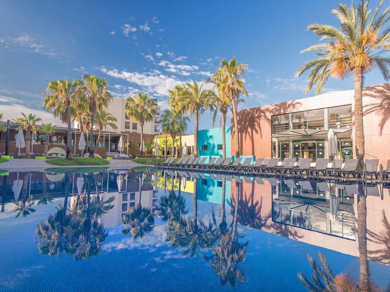 Hotel mit Pool, Palmen und klarem Himmel, Gebäude in Weiß, Blau und Orange reflektiert im Wasser.