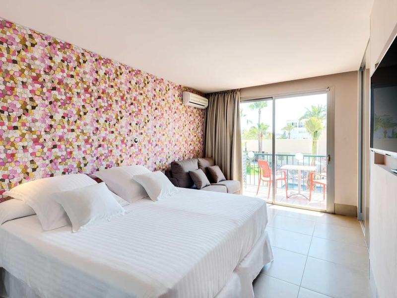 Helles Hotelzimmer mit drei Einzelbetten, bunter Blumenwand und Balkon mit Aussicht.