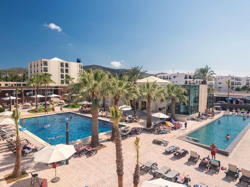 Hotelanlage mit zwei großen rechteckigen Schwimmbecken, Palmen und Liegestühlen unter blauem Himmel.