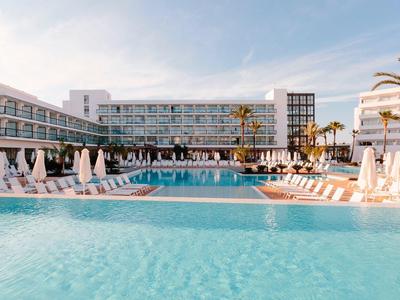 Modern hotel with large pool and sun loungers under a partly cloudy sky.