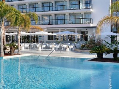 Moderner Hotelpool mit klarem Wasser und Liegestühlen unter Sonnenschirmen neben dem Gebäude.