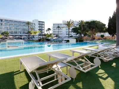 Hotelgarten mit Pool und Liegestühlen unter Palmen an sonnigem Tag.