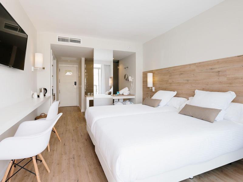 Bright hotel room with twin beds, white decor, wooden floor, and a wall-mounted TV.