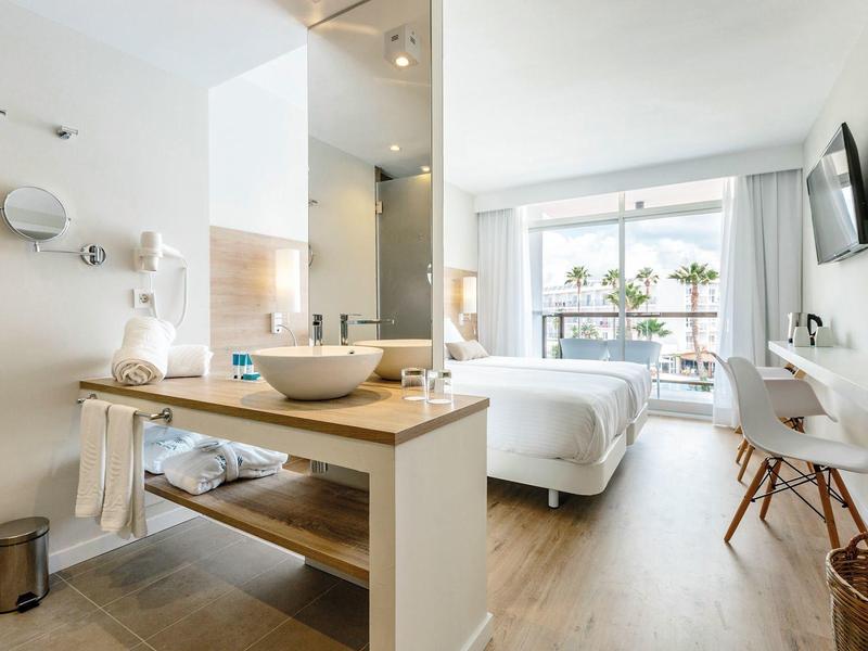 Bright hotel room with double bed, desk, chair, and large window overlooking palm trees.