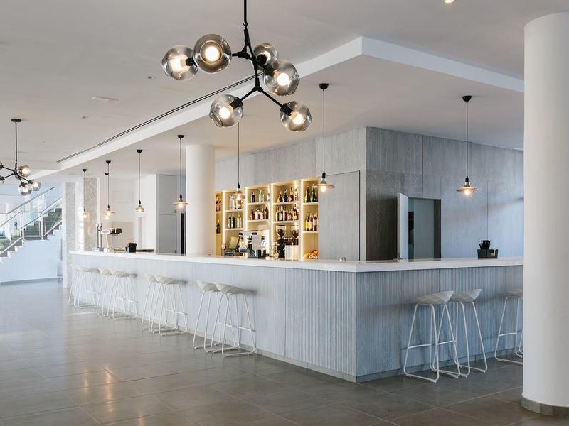 Modern hotel lobby with a long white bar counter, stools, and contemporary lighting fixtures.