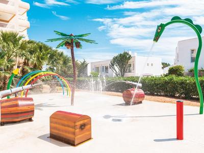 Kinderspielplatz mit Wasserspielen, Palmen, bunten Bögen und hellen Sandflächen unter blauem Himmel.