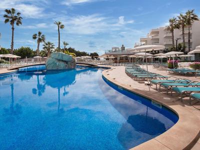 Großer Außenpool mit Liegestühlen und Sonnenschirmen in Hotelanlage unter blauem Himmel.