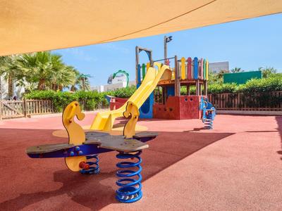 Kinderspielplatz mit bunter Rutsche und Federspielgerät auf rotem Bodenbelag unter blauem Himmel.