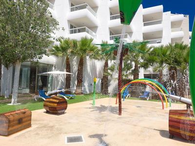 Kinderspielplatz mit Sand, Rutschen und Bänken vor einem weißen Hotelgebäude und Palmen.