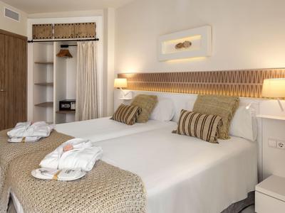 Ein hell eingerichtetes Hotelzimmer mit zwei Betten, dekorativen Kissen und offener Garderobe.