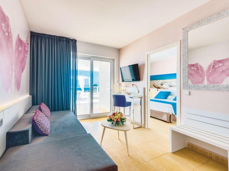 Helles Zimmer mit Sofa, kleinem Tisch, Balkon, blauem Stuhl und Doppelbett mit maritimen Bildern.