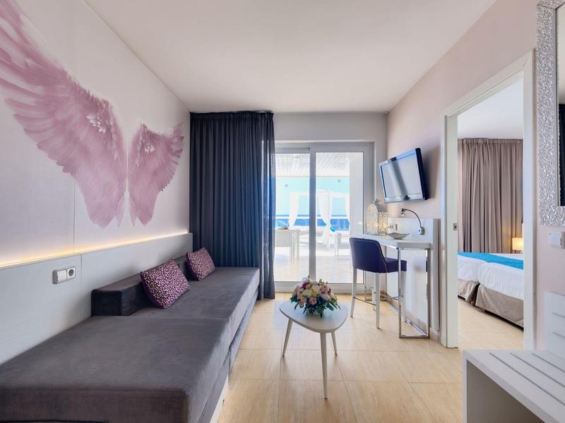 Helles Wohnzimmer mit grauem Sofa, Vögel-Wandbild, kleinem Tisch und Balkon mit blauem Himmel.